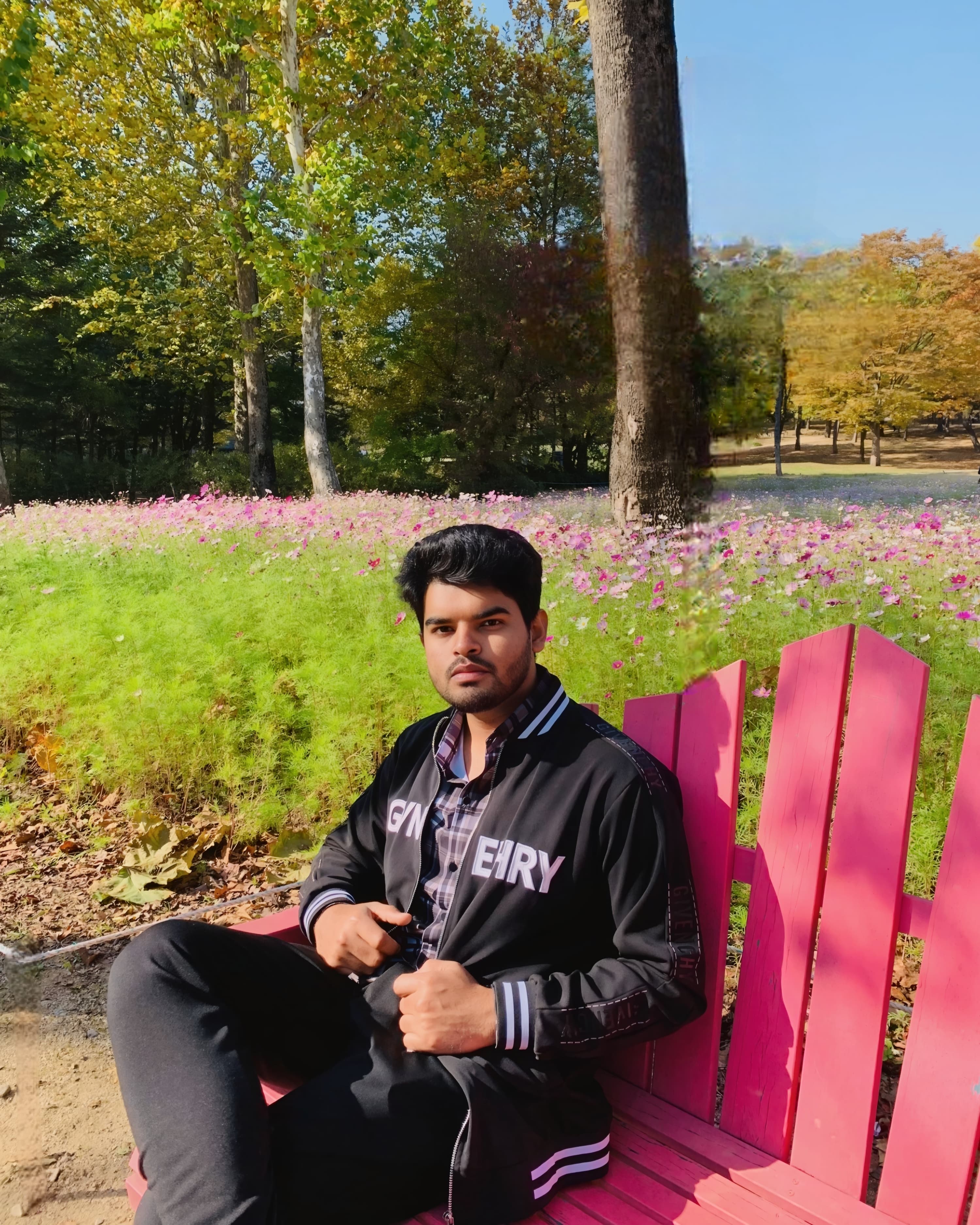 Shawkat Hossain Maruf aka shawkath646 sitting on a red bench in a flower field with trees showing early autumn colors in the background, enjoying the sunshine.