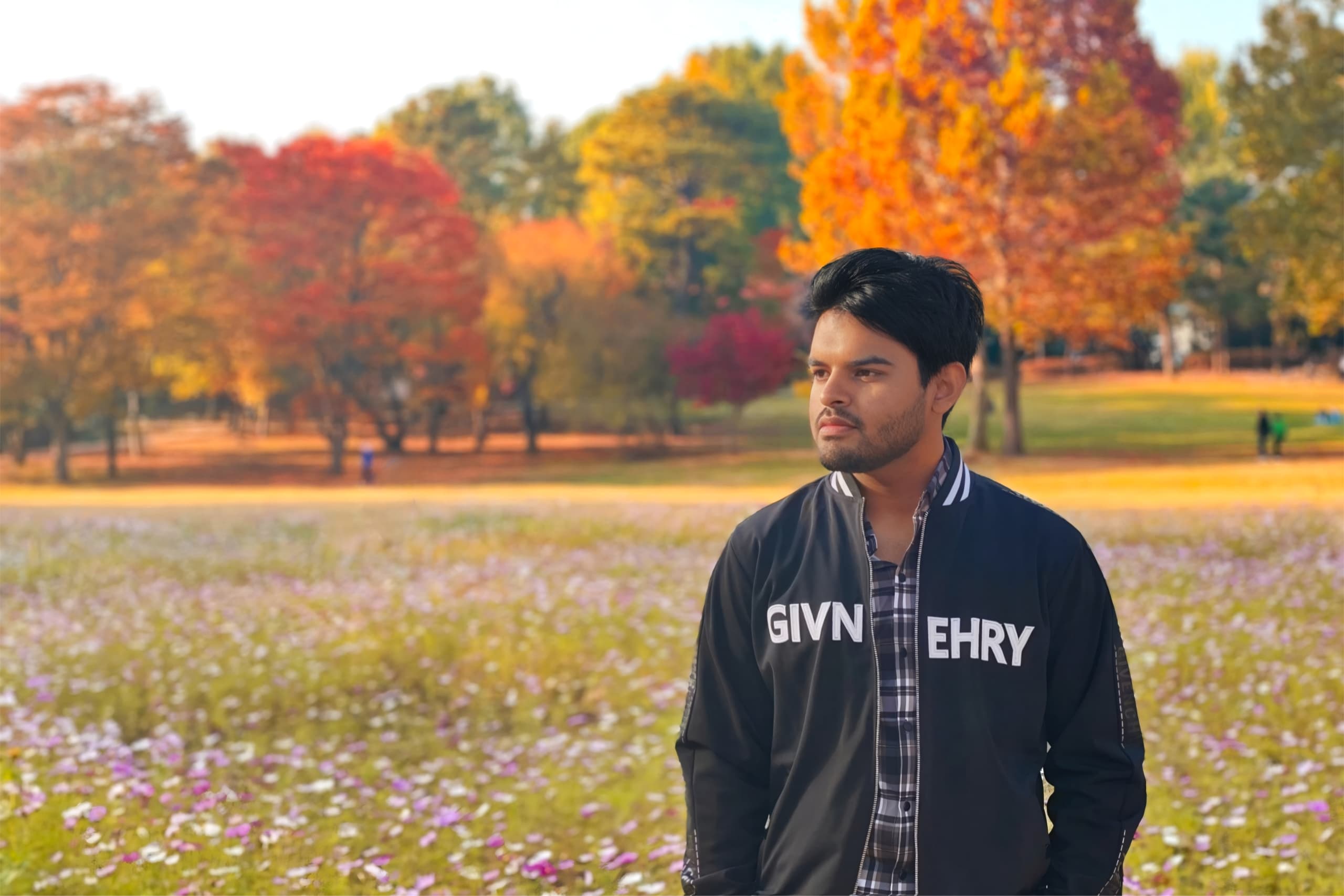 Thoughtful shawkath646 in a checkered shirt and black jacket in a meadow of autumn wildflowers, with fall trees in the distance